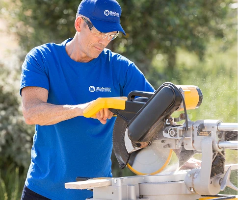 Technician in a Handyman Connection uniform uses a yellow miter saw to cut a piece of wood outdoors.