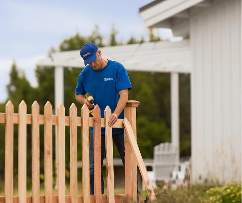 Technician in a blue Handyman Connection uniform uses a cordless drill to fasten boards on a wooden picket fence outdoors near a white building.